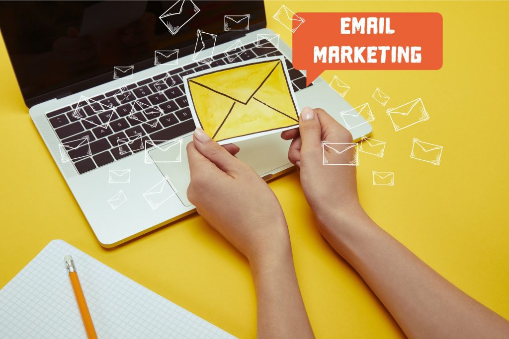 cropped image of woman holding envelope sign near laptop with"email marketing" lettering and email